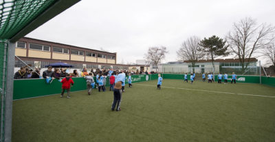 Zu sehen ist das Mini-Sportfeld mit Kunstrasen auf dem Schulhof der Grundschule Bous mit Fußball spielenden Kindern.