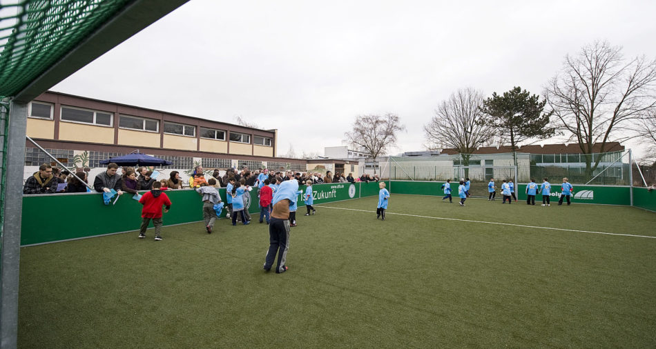 Zu sehen ist das Mini-Sportfeld mit Kunstrasen auf dem Schulhof der Grundschule Bous mit Fußball spielenden Kindern.