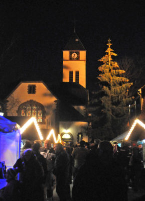zu sehen sind beleuchtete Weihnachtshütten mit vielen Menschen davor und im Hintergrund die beleuchtete und von innen strahlende evangelische Kirche