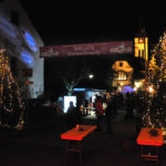 zu sehen ist der beleuchtete Eingang am Weihnachtsmarkt mit zwei Tannenbaeumen links und rechts und der im Hintergrund beleuchteten evangelischen Kirche sowie LED Spots der Bayern Bazis Bous die auf eine Hauswand das Bayern Logo projizieren 