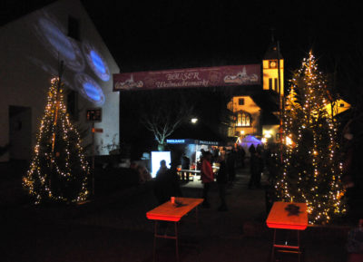 zu sehen ist der beleuchtete Eingang am Weihnachtsmarkt mit zwei Tannenbaeumen links und rechts und der im Hintergrund beleuchteten evangelischen Kirche sowie LED Spots der Bayern Bazis Bous die auf eine Hauswand das Bayern Logo projizieren